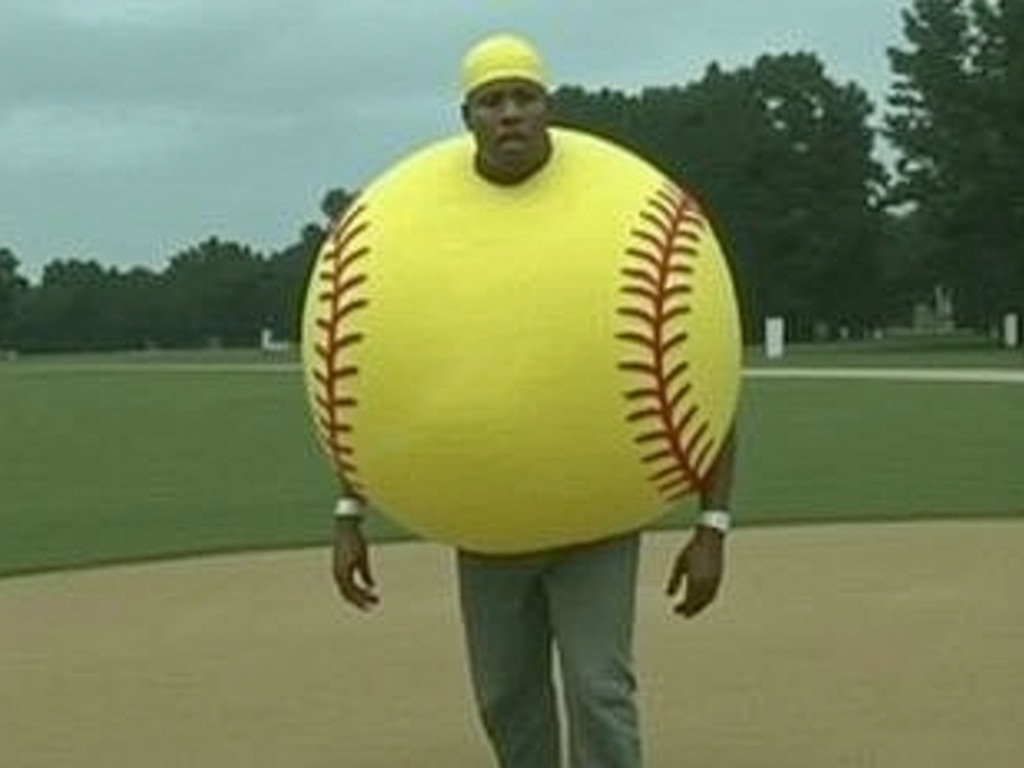 Man standing on a baseball infield inside a giant round yellow softball costume, wearing a deadpan expression
